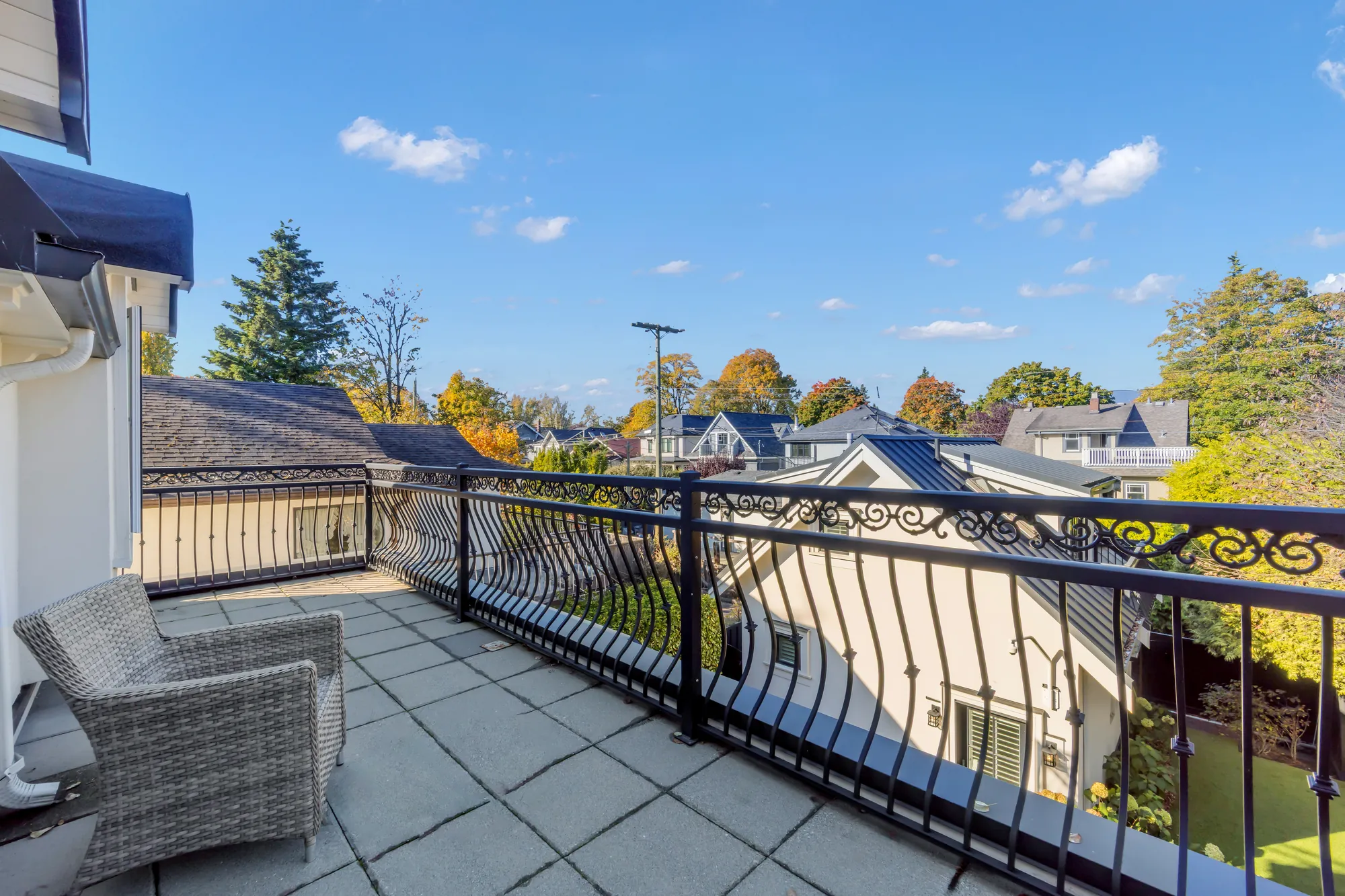 The balcony off the primary bedroom: stone pavers, a curved wrought-iron railing, wicker chairs, roofs and autumn trees beyond