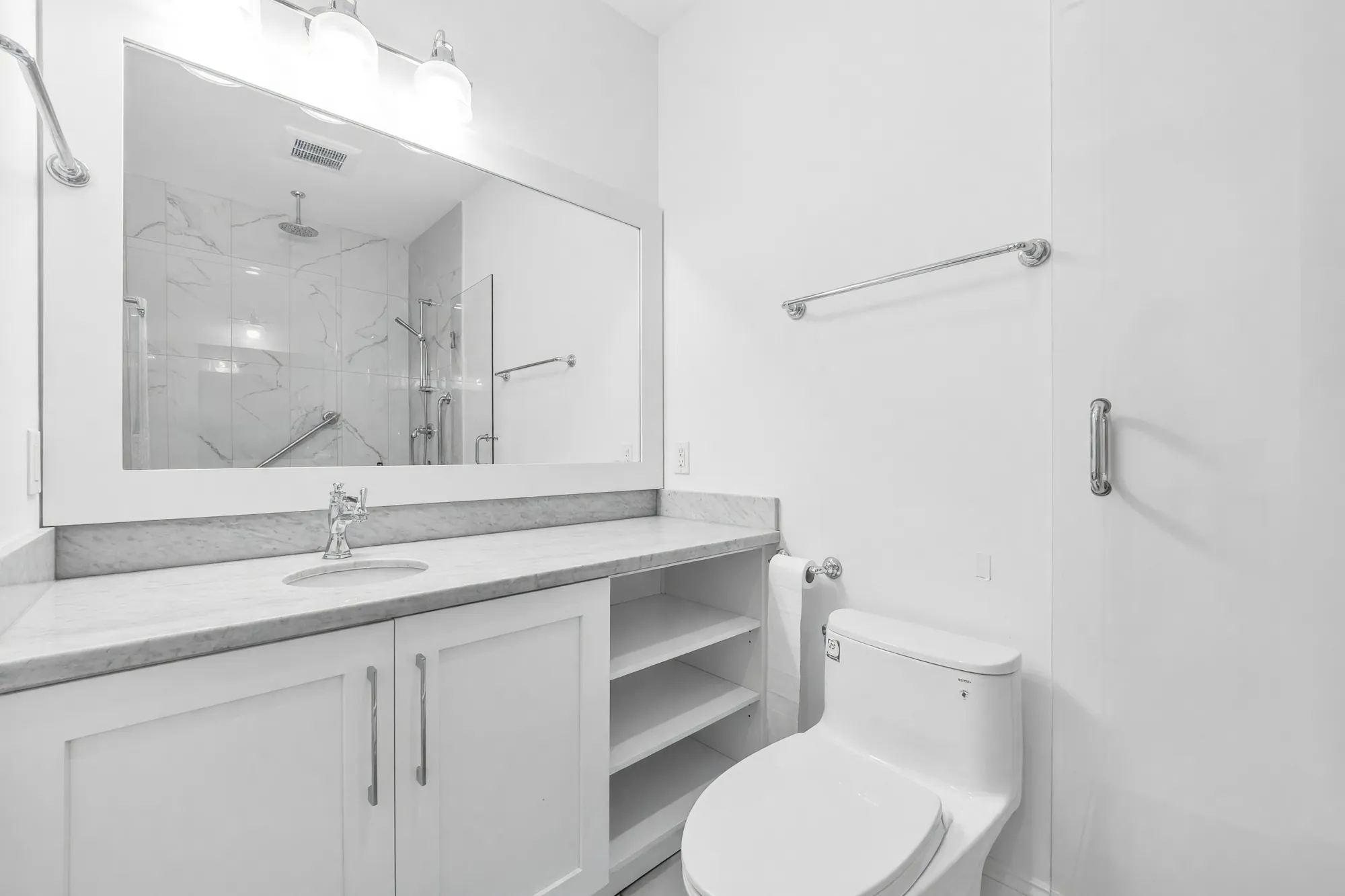 The lower bathroom: a white vanity with a grey stone top and open shelves, a glass shower reflected in the mirror, a toilet
