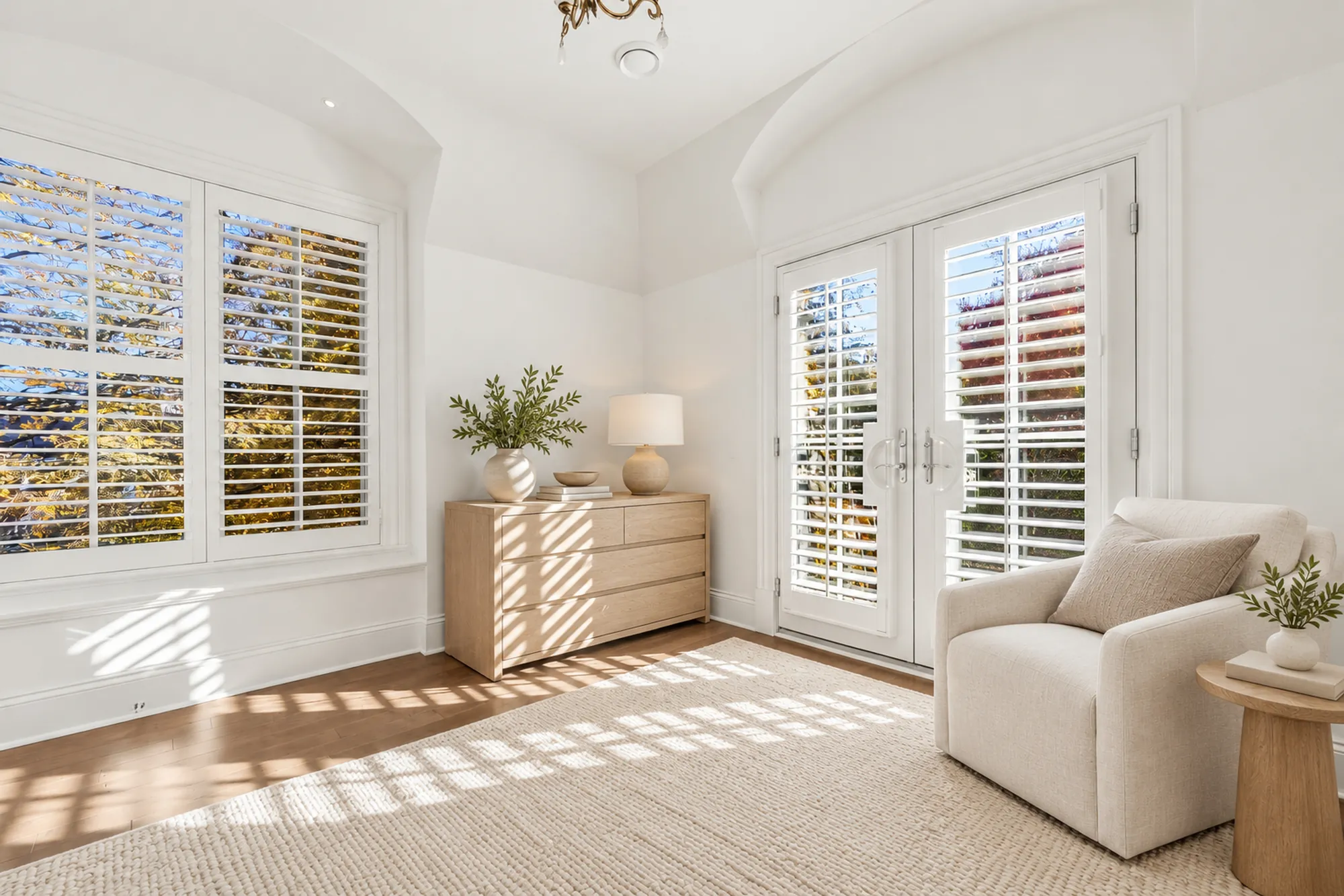 An upstairs bedroom virtually staged: a pale wood dresser and an armchair on a jute rug, a wide shuttered window and French doors, sunlight on the floor