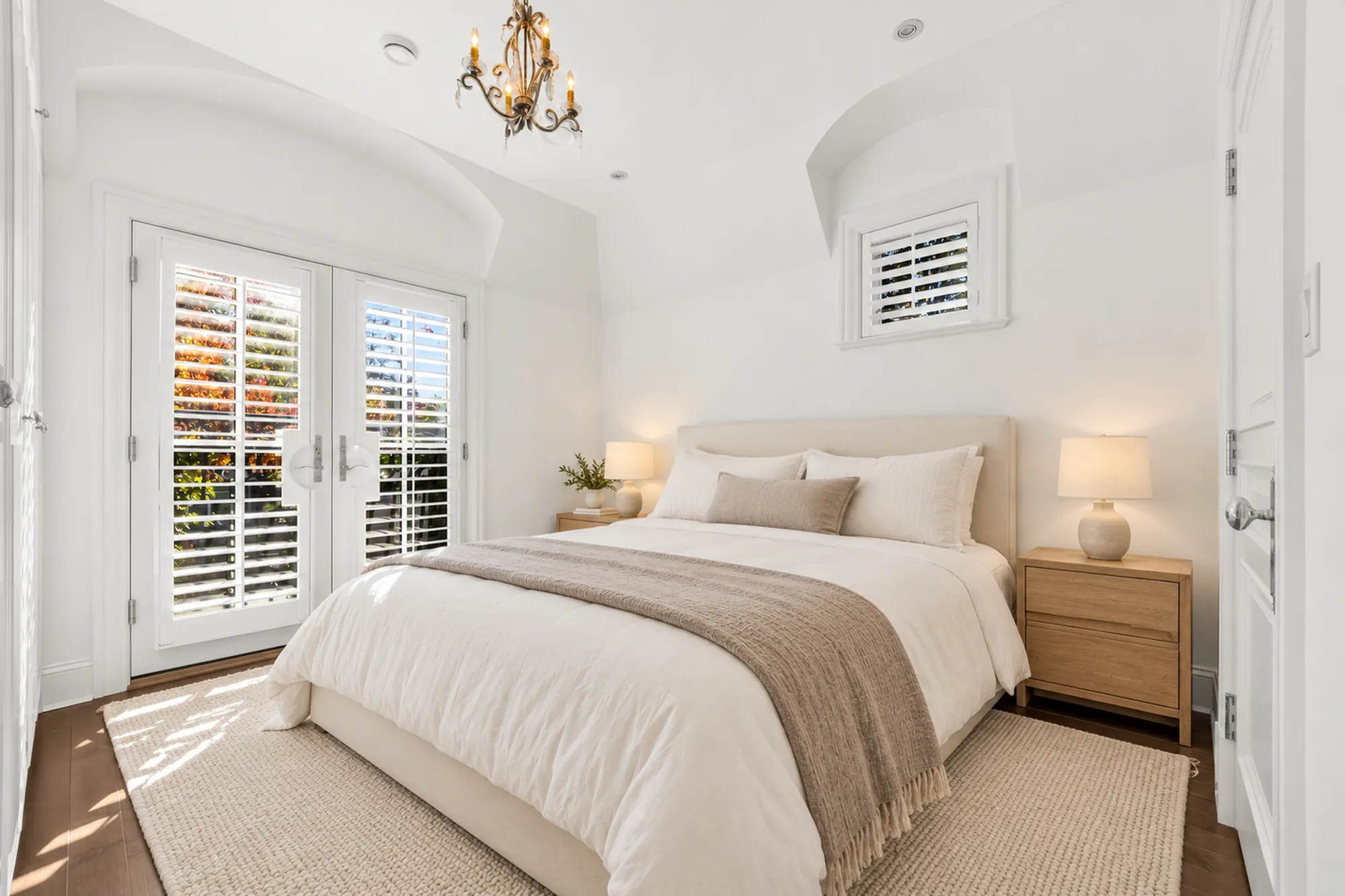 An upstairs bedroom virtually staged: a cream bed with a taupe throw, night tables and lamps, a chandelier, French doors and a small shuttered window