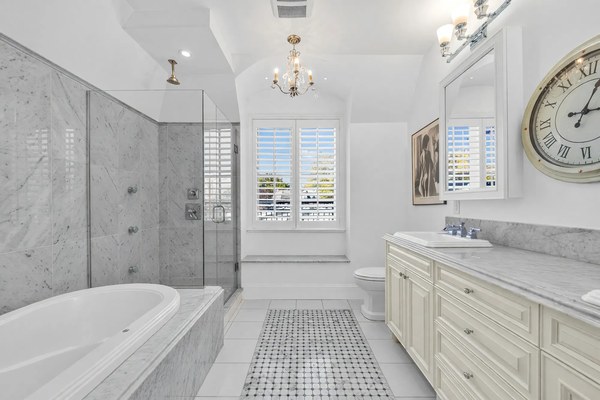 The primary bathroom: a soaking tub, a marble shower with a glass door, a long cream vanity with a marble top, a chandelier and shuttered windows