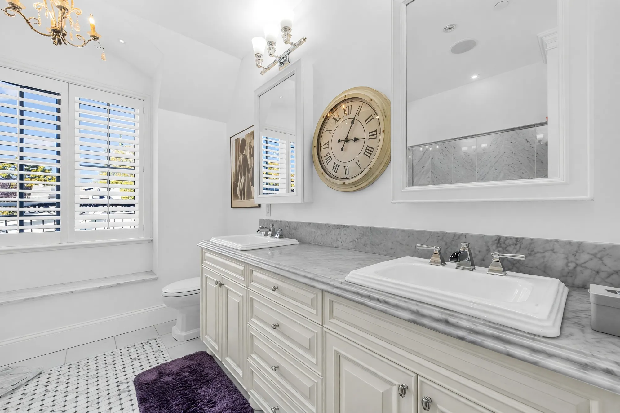 The double vanity in the primary bathroom: two sinks on a grey marble top, a large clock and mirrors, a shuttered window