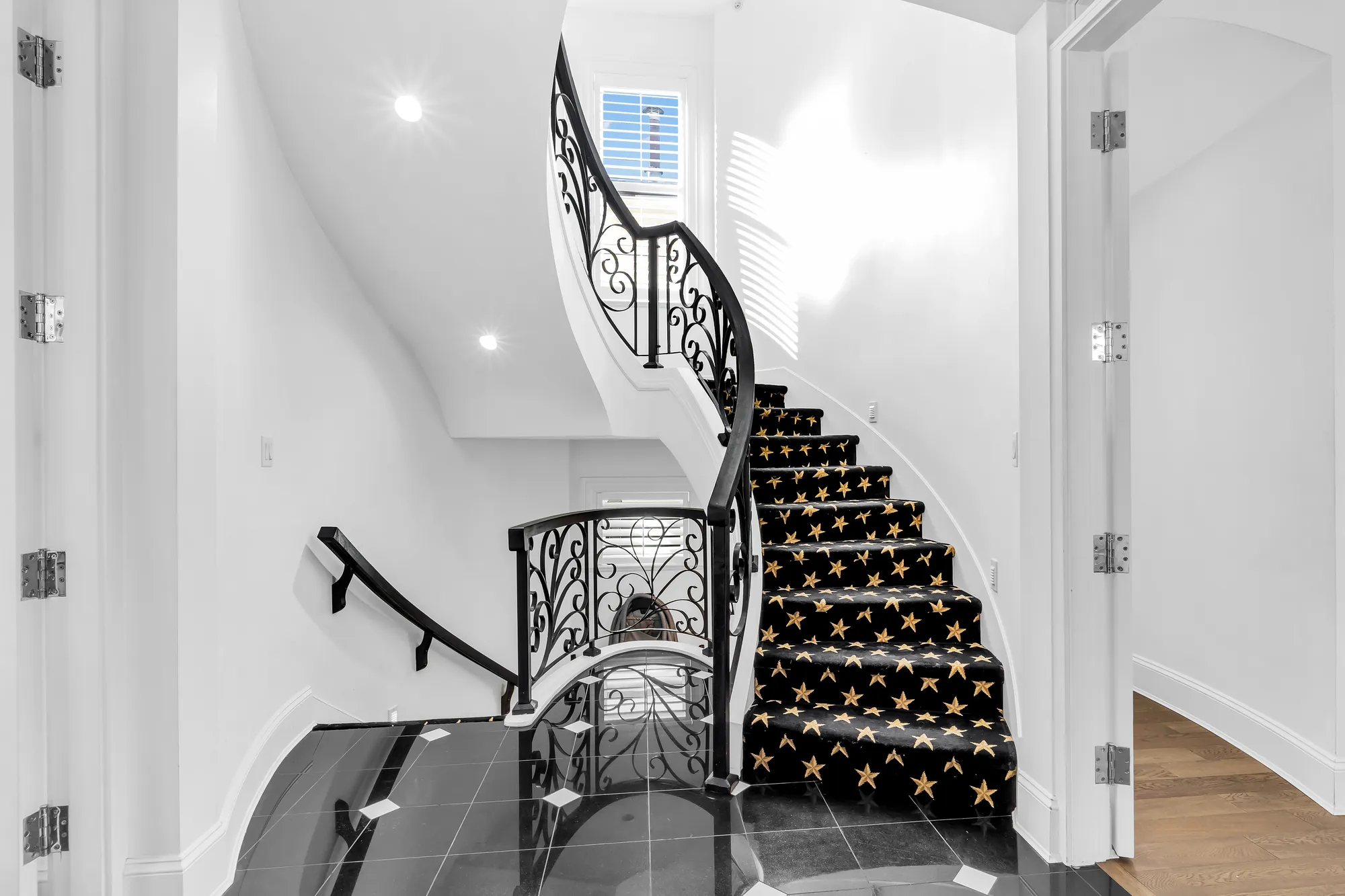 The foyer: a curved staircase with a wrought-iron railing and a carpeted run, black stone tiles, white walls, the stair down to the lower floor