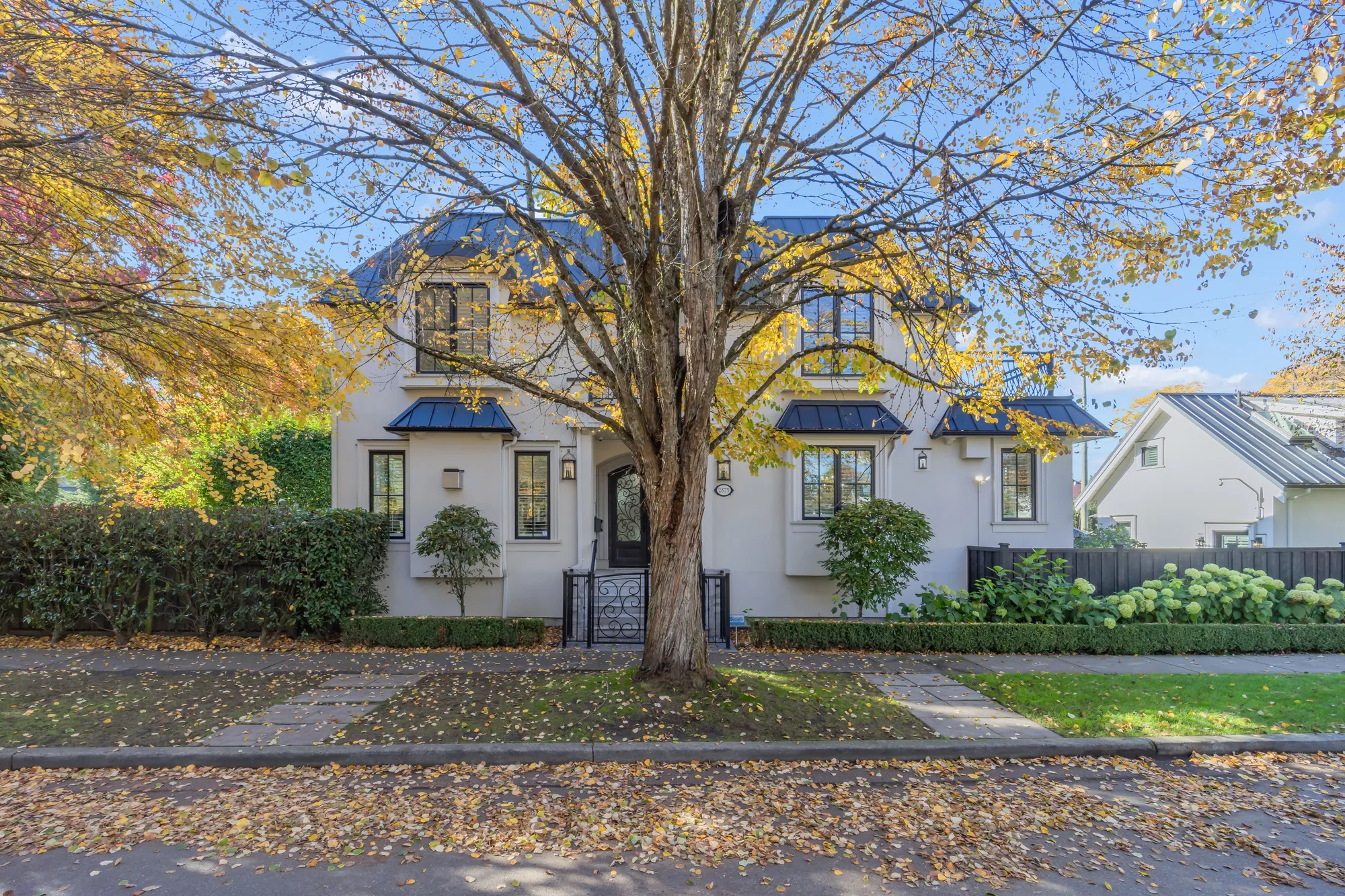 The front of the house in autumn: cream stucco, black metal awnings over the windows, an elm with gold leaves on the lawn, a hedge and an iron gate to the front steps