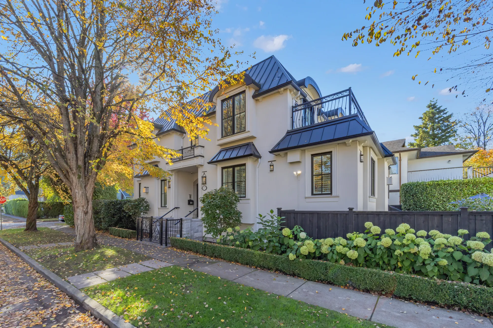 The front of the house at an angle: the bay windows under black metal awnings, a balcony on the upper floor, hydrangeas behind a low hedge