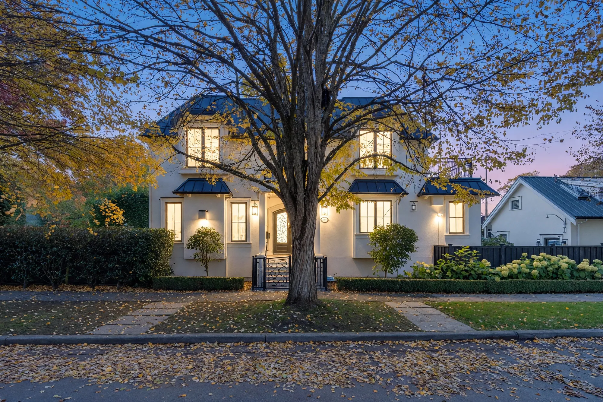 2875 Trimble Street at dusk: a cream stucco house with black metal awnings, the windows lit, an elm with the last of its gold leaves on the lawn