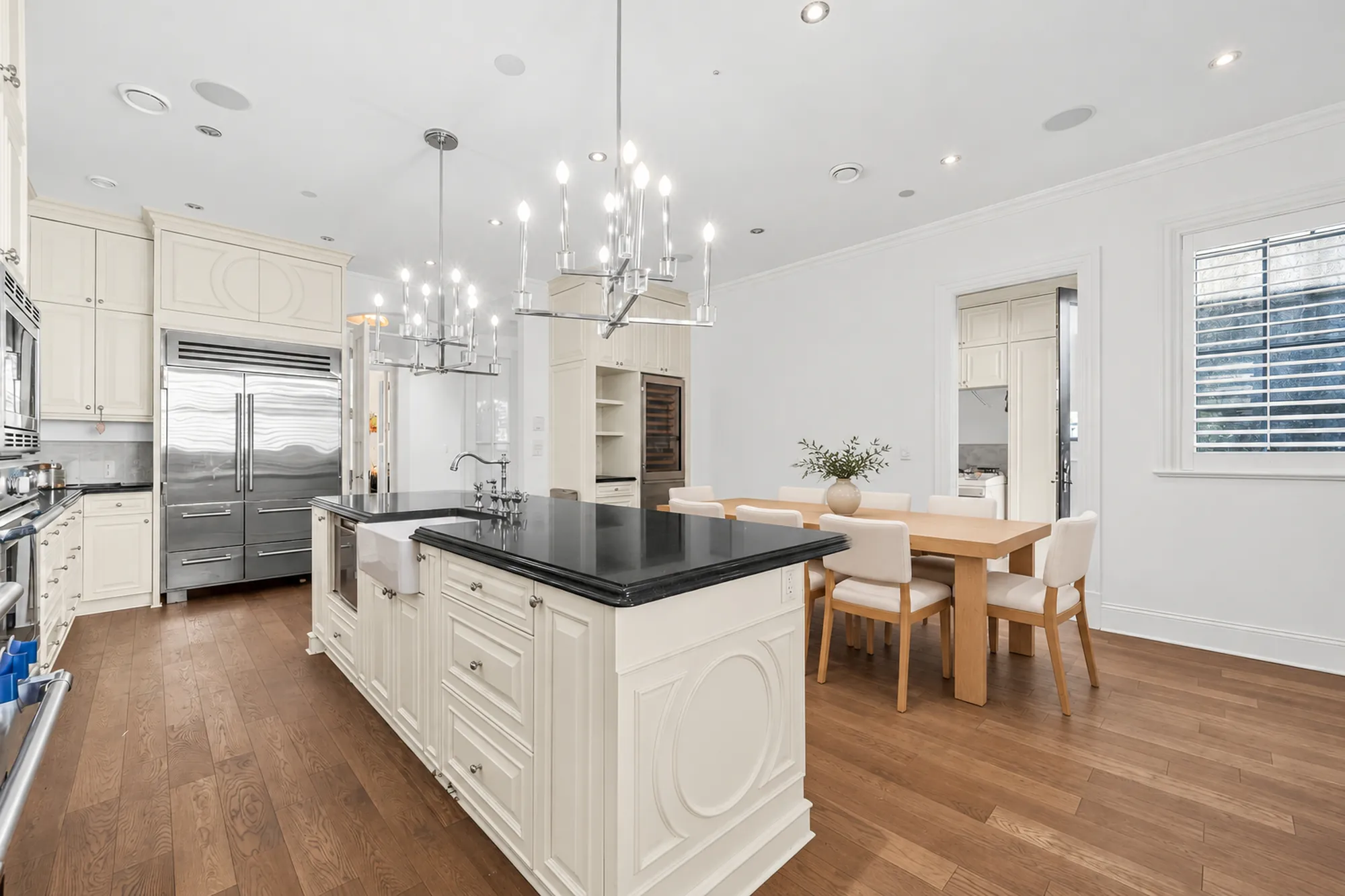 The kitchen virtually staged: a long island with a farmhouse sink, a built-in refrigerator, chandeliers, the dining table by the windows