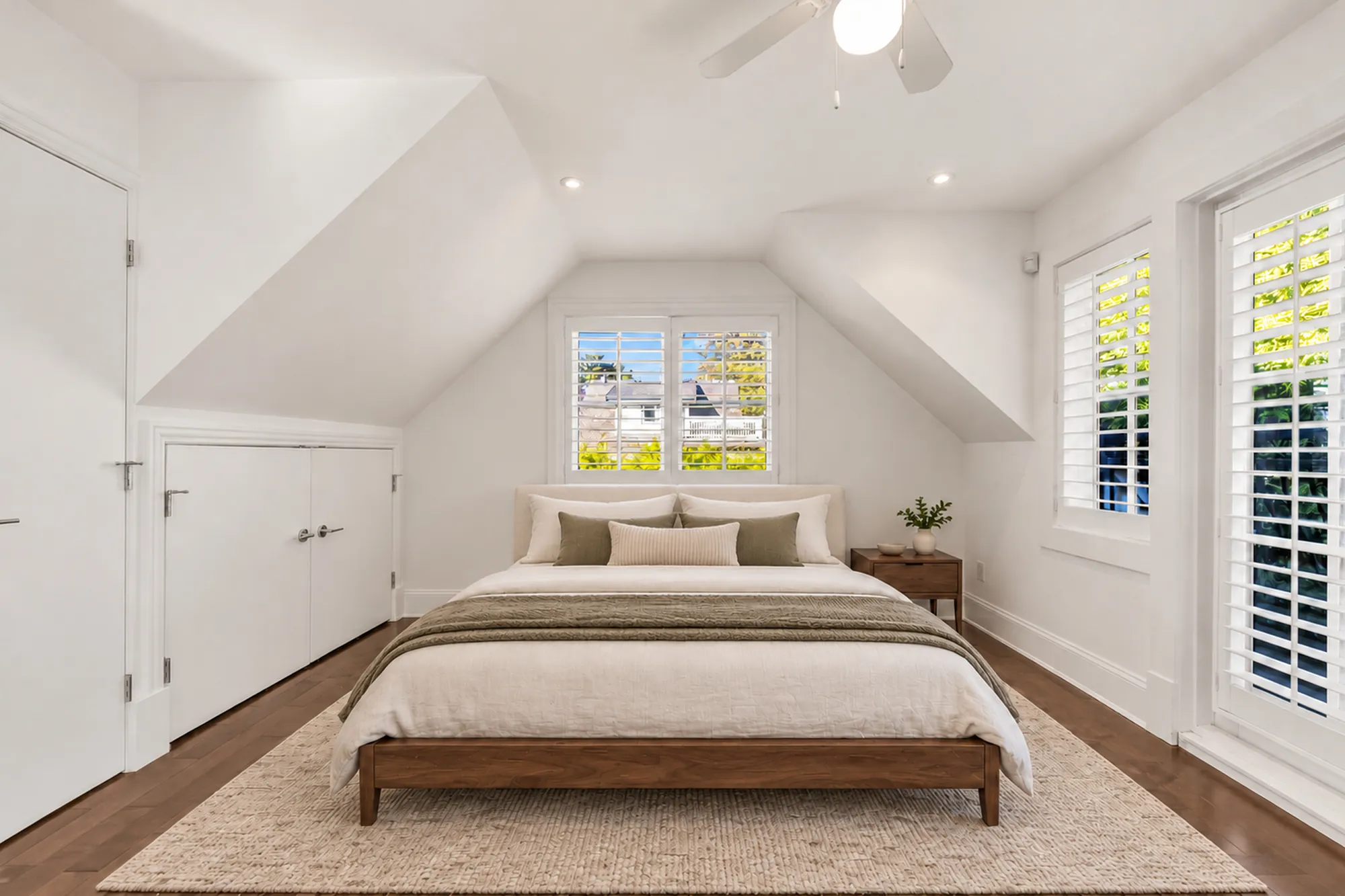 The laneway bedroom virtually staged: a wood bed under the gable window, sloped ceilings, a jute rug, French doors with shutters