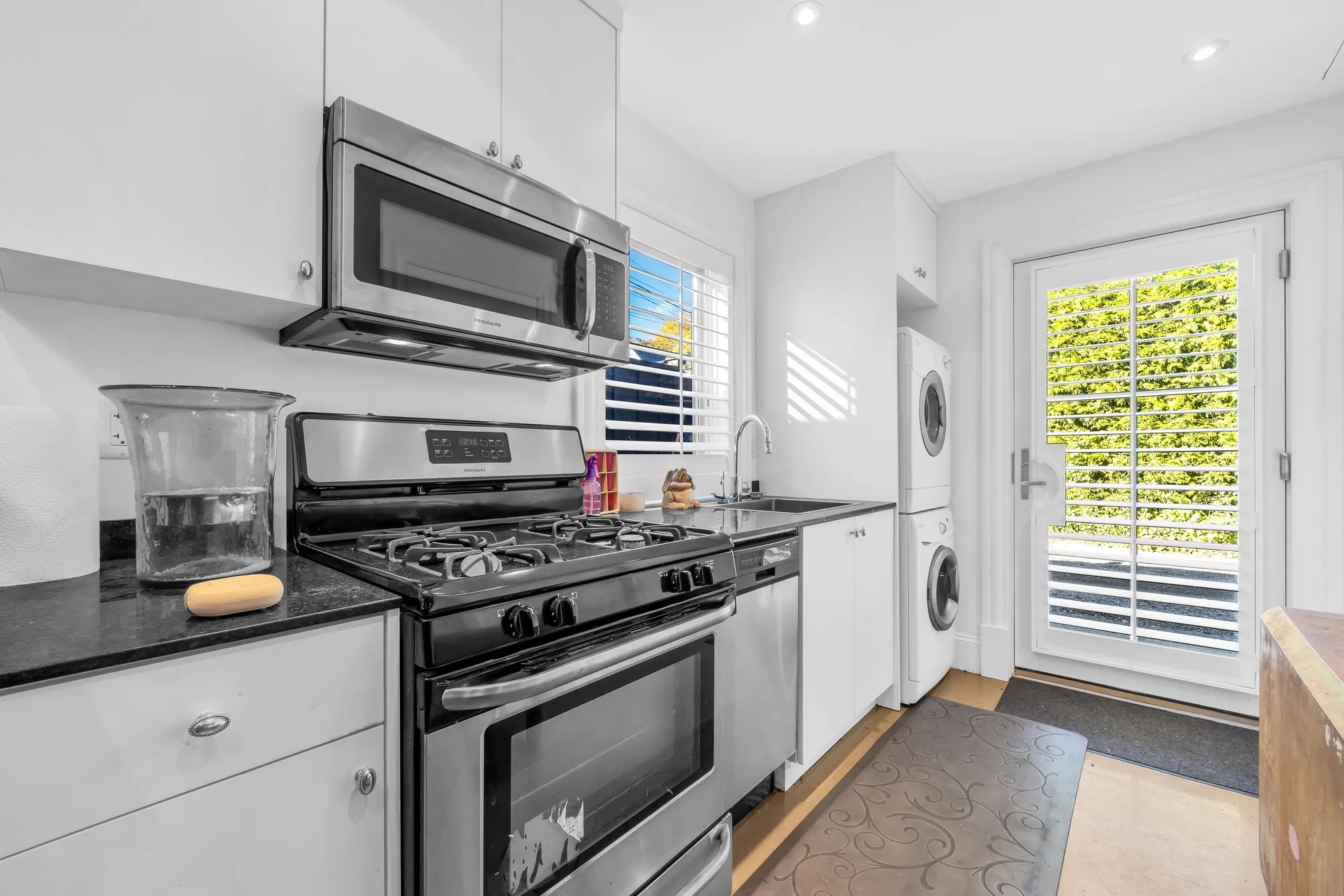The laneway kitchen: white cabinets, a gas range and microwave, a black counter and sink, a stacked washer and dryer, a glass door to the garden