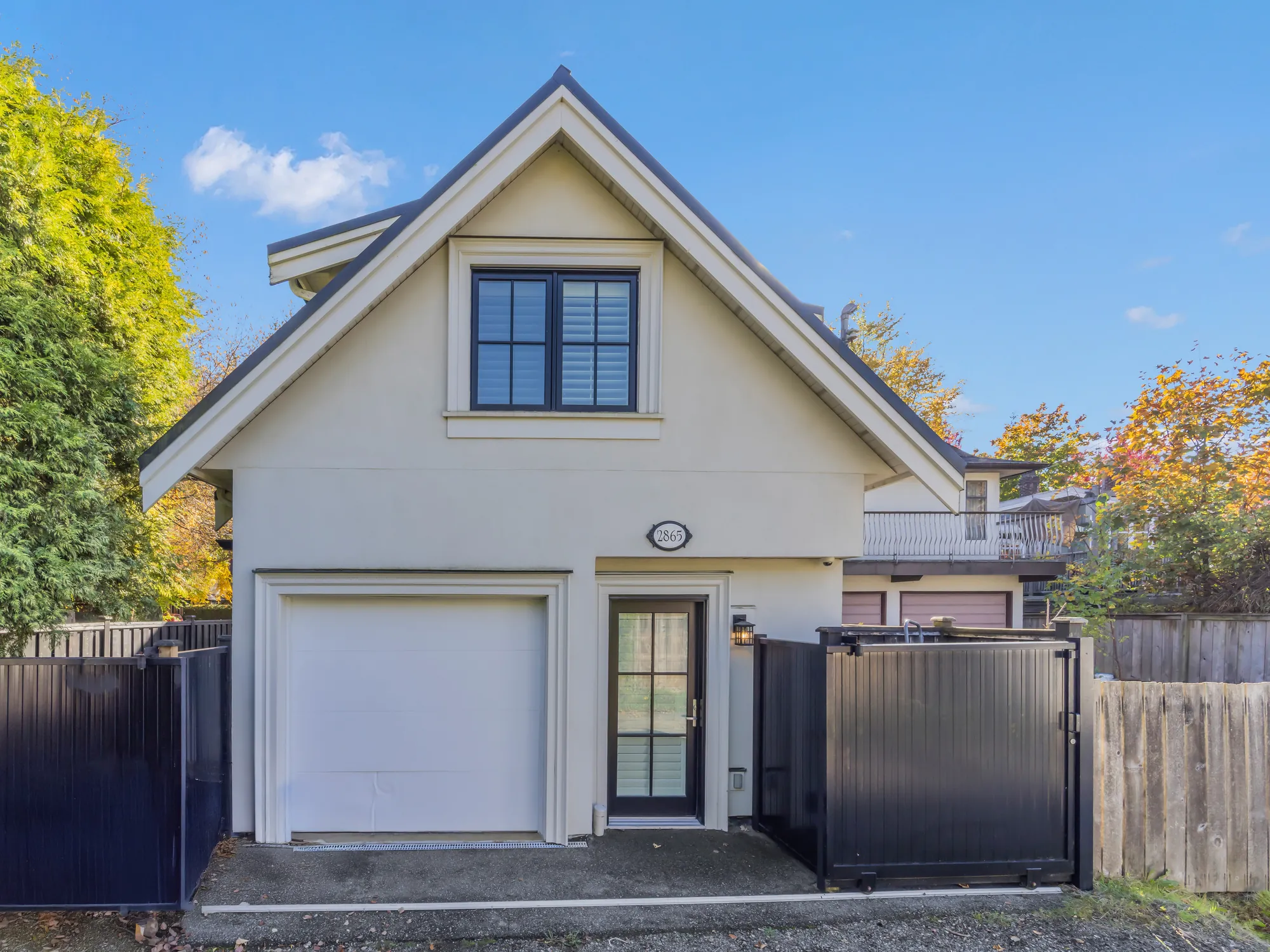 The laneway house from the lane: a gabled cream house with a single garage door, a glass door with a light above, black gates on either side