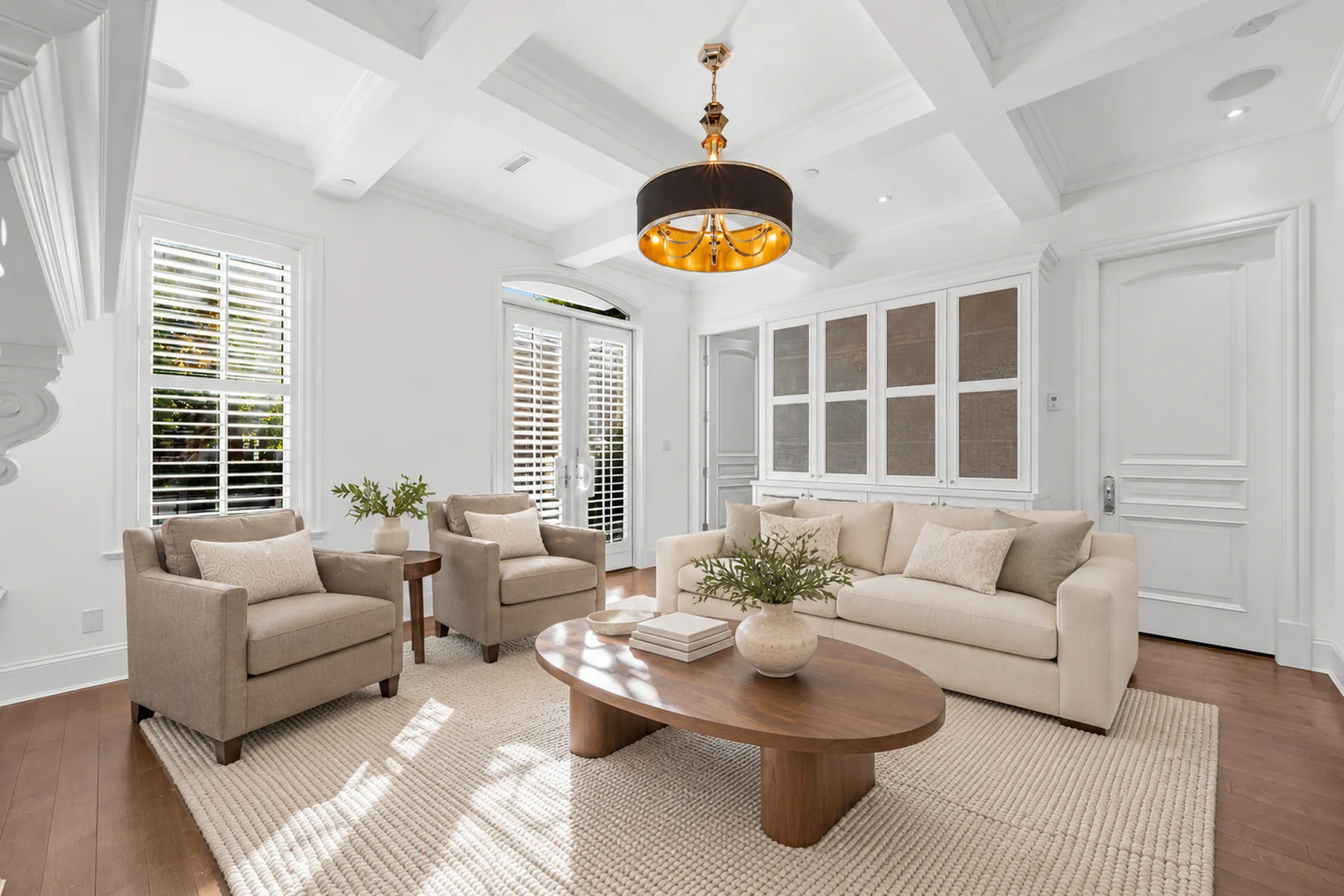 The living room from the fireplace end, virtually staged: two armchairs and a sofa on a jute rug, the coffered ceiling and a drum pendant, French doors and a built-in cabinet