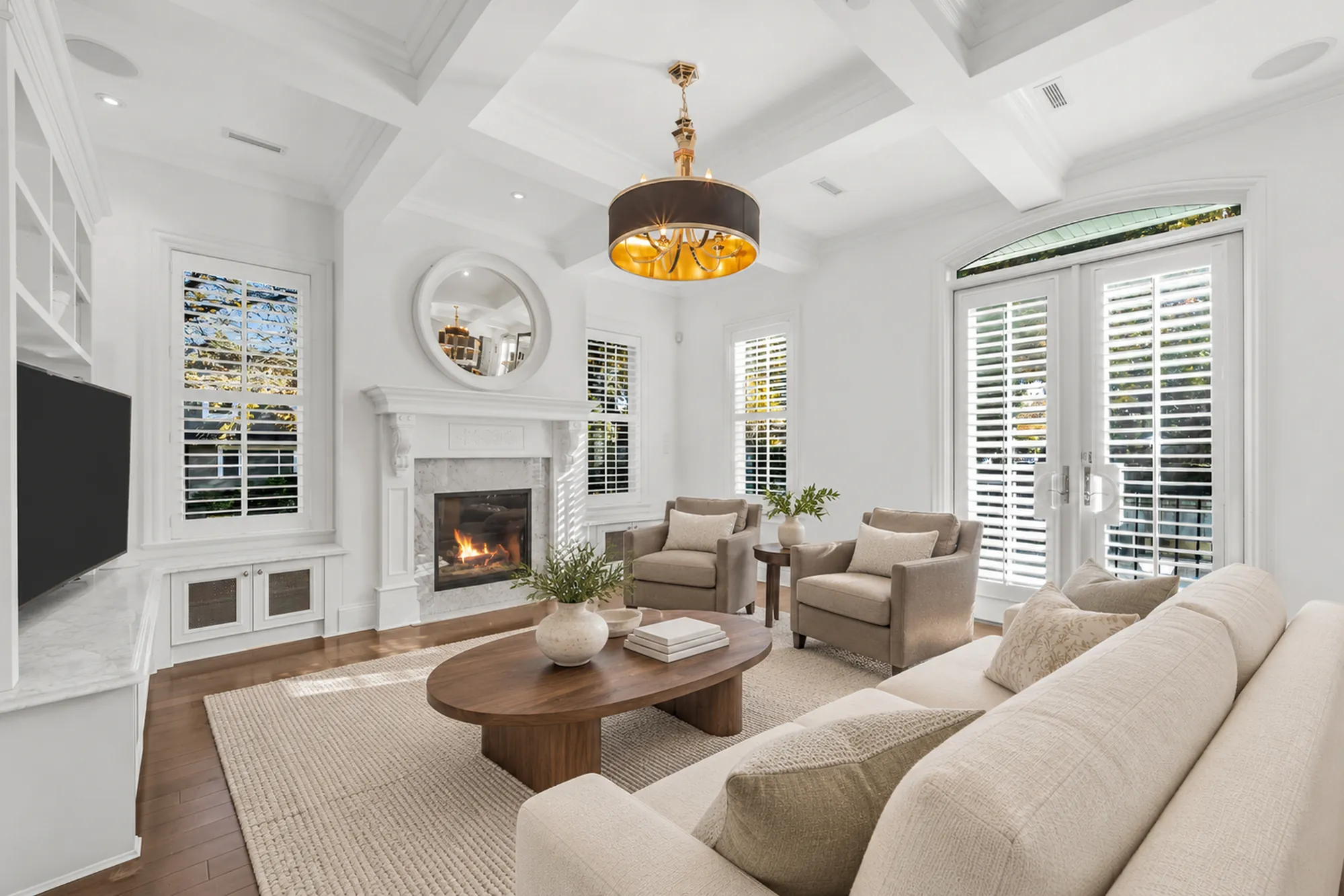 The living room virtually staged: a cream sofa and two armchairs around an oval wood coffee table on a jute rug, a gas fireplace under a round mirror, a coffered ceiling, French doors with shutters