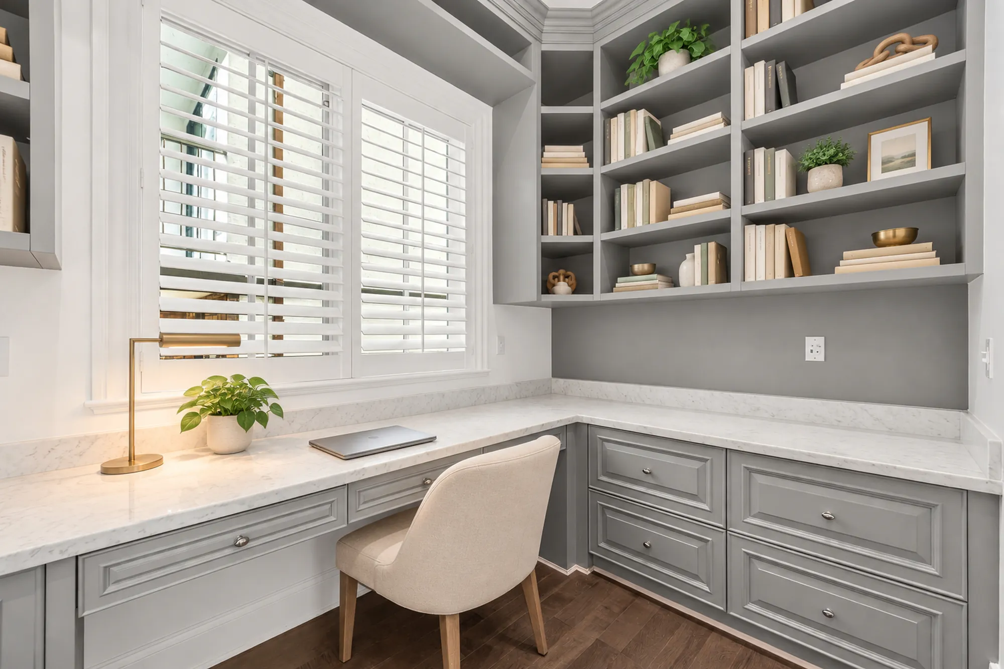 The office virtually staged: a built-in desk with grey cabinets and a marble top, open shelves above, a chair, a brass lamp, shuttered windows