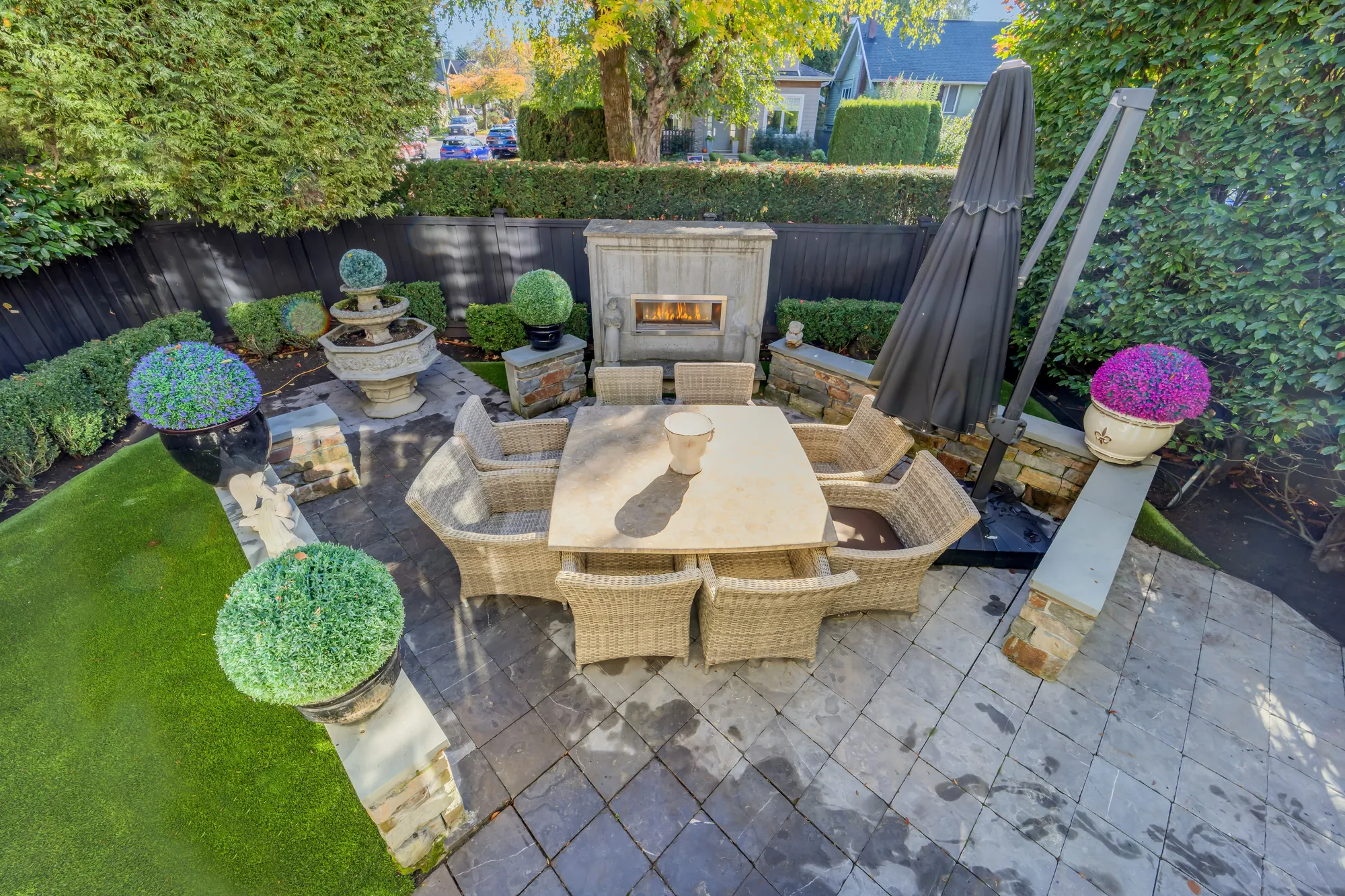 The patio from above: an outdoor dining table and wicker chairs on stone paving, a gas fireplace in a stone surround at the end, clipped topiary