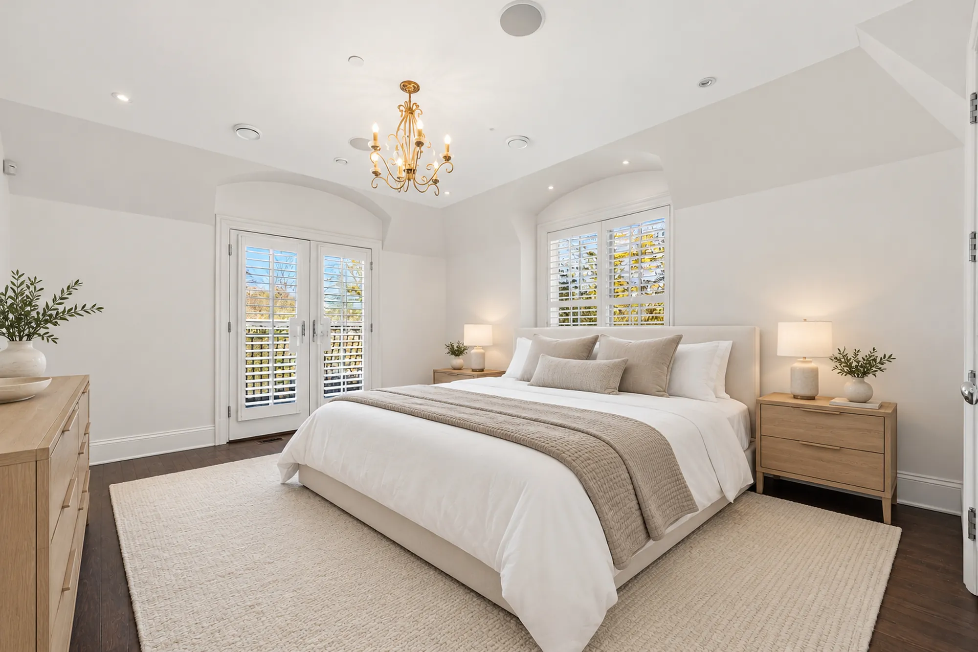 The primary bedroom virtually staged: a cream upholstered bed with linen bedding, wood night tables and lamps, a chandelier, French doors and a shuttered window