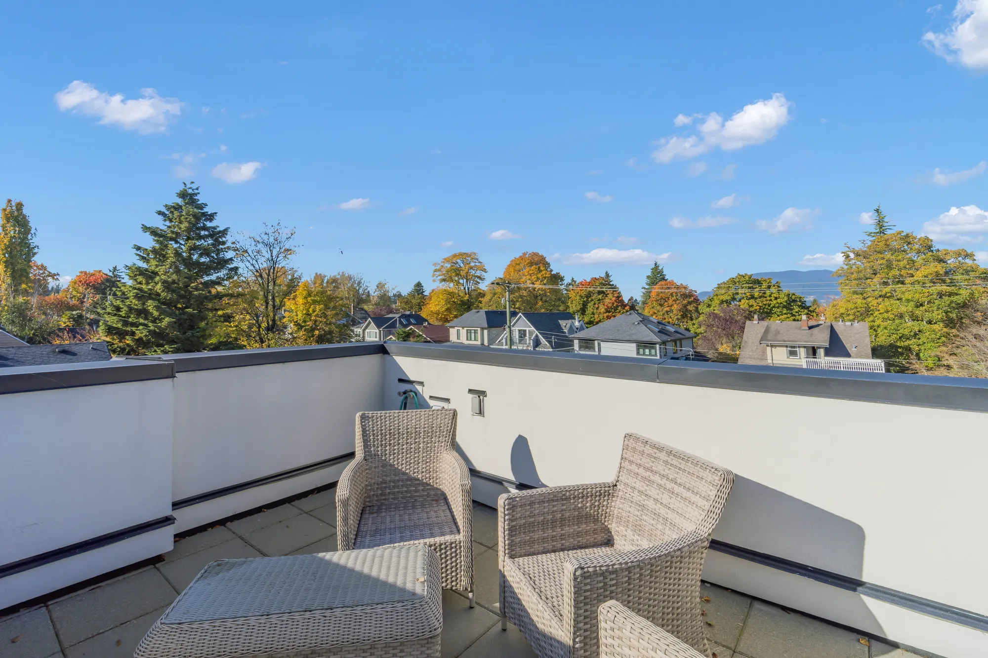 The roof deck: stone pavers inside a white parapet, wicker armchairs and a table, treetops and the North Shore mountains on the horizon