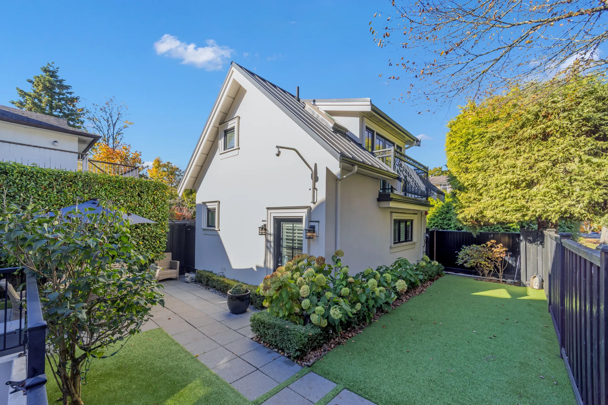 A side garden: a paved path between a lawn and a bed of hydrangeas, the laneway house at the back, a black fence